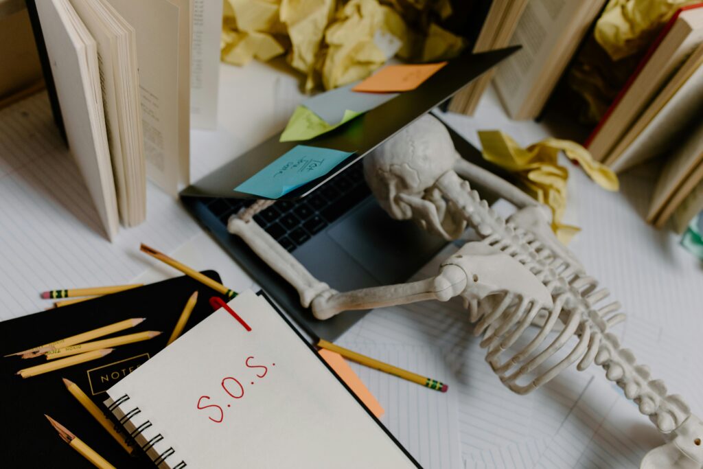 pexels-photo-8386709-8386709 Dramatic still life of a skeleton overwhelmed at an untidy office desk with S.O.S. note.